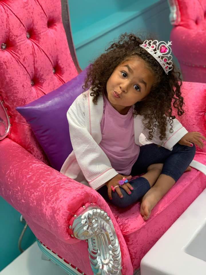 A little girl sitting in a pink chair wearing a tiara.